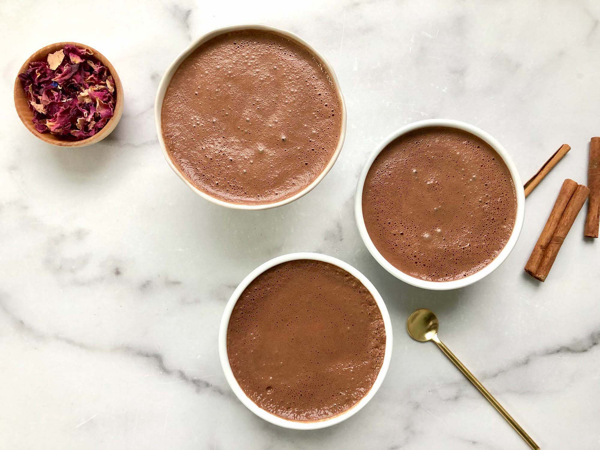 Three cups of ceremonial cacao with cinnamon sticks and a bowl of rose petals on a marble counter