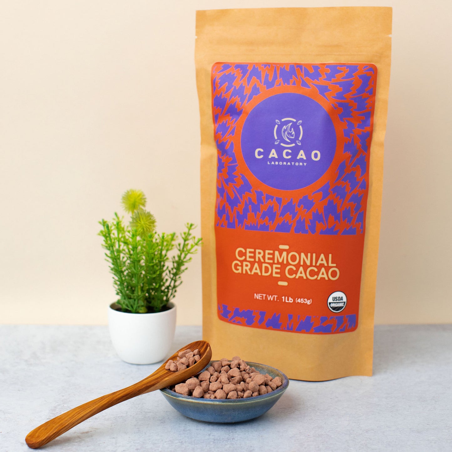 A stand-up pouch of Cacao Laboratory Ceremonial Grade Cacao sits on a light gray surface with a small bowl of cacao nibs and a wooden spoon in front, and a small potted green plant to the left. The pouch is kraft brown with a large purple and orange label.