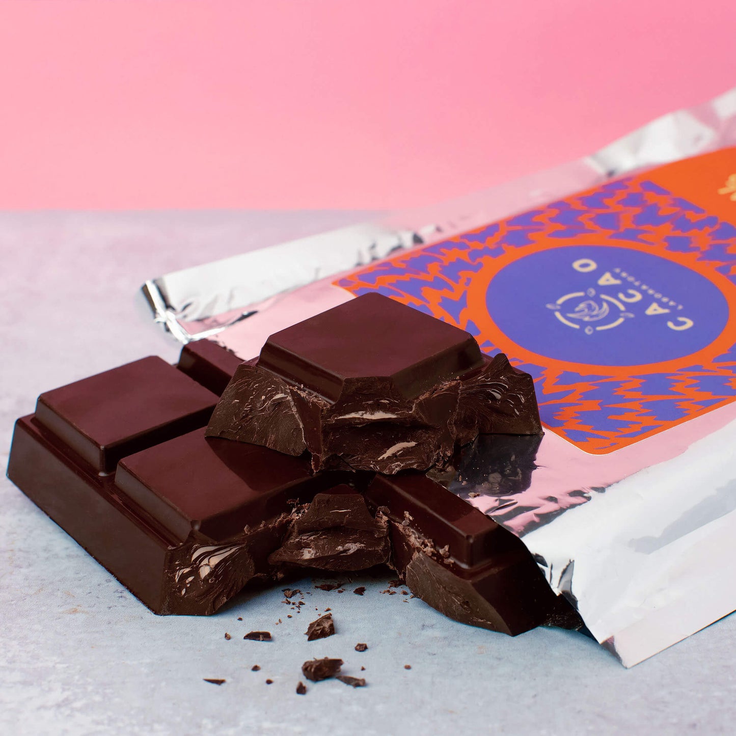 A close-up of broken Cacao Laboratory 100% cacao bar placed in front of a partially unwrapped foil package with a pink, orange, and blue label. The background is a soft pink upper area and a light gray surface below.
