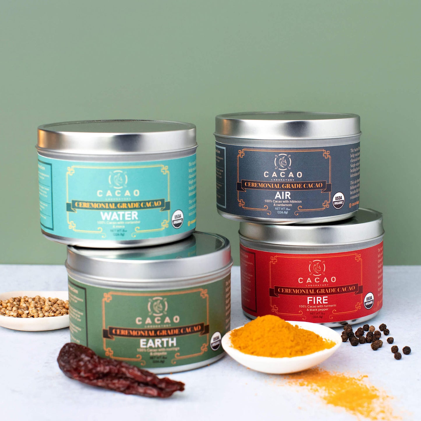 Four ceremonial cacao by Cacao Laboratory tins labeled 'Water', 'Air', 'Fire', and 'Earth' with cacao and ingredients on a light green background.