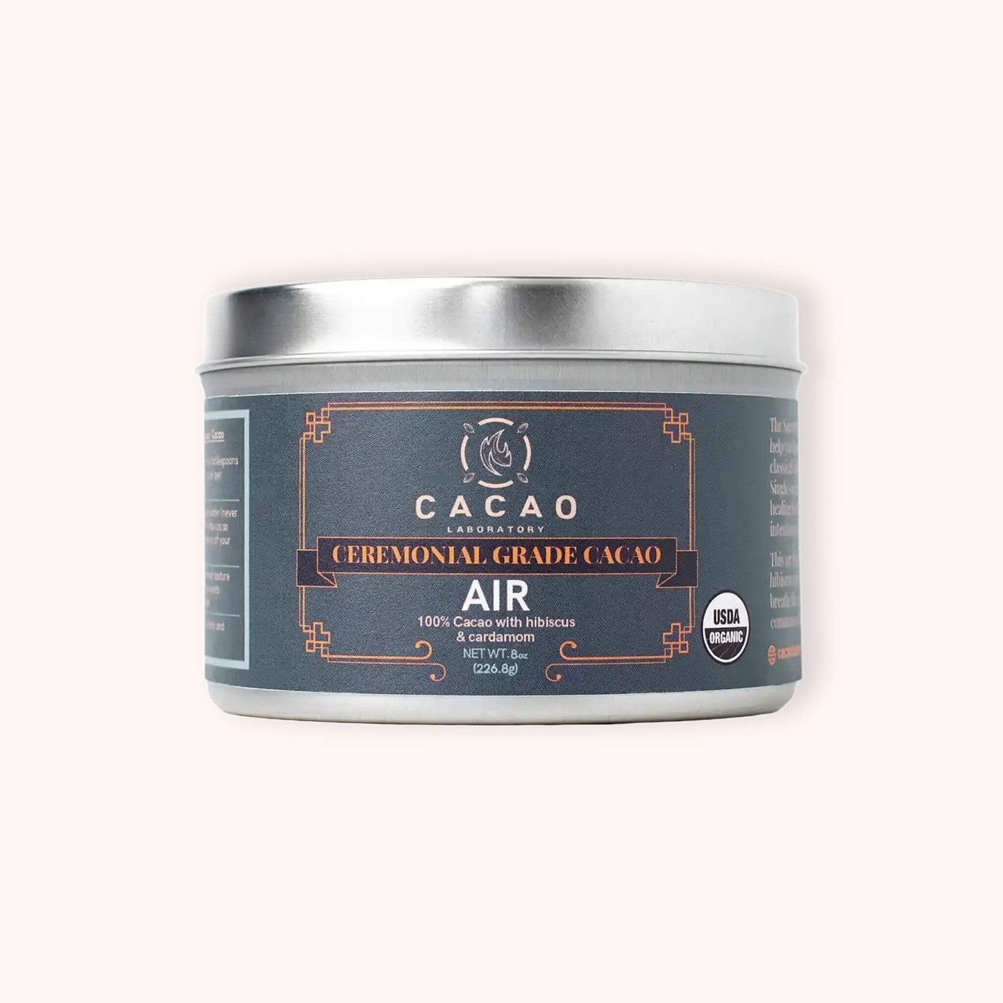 A silver metal tin of Cacao Laboratory 'Air' ceremonial grade cacao, featuring a dark slate blue label with orange accents, set against a solid pale pink background.
