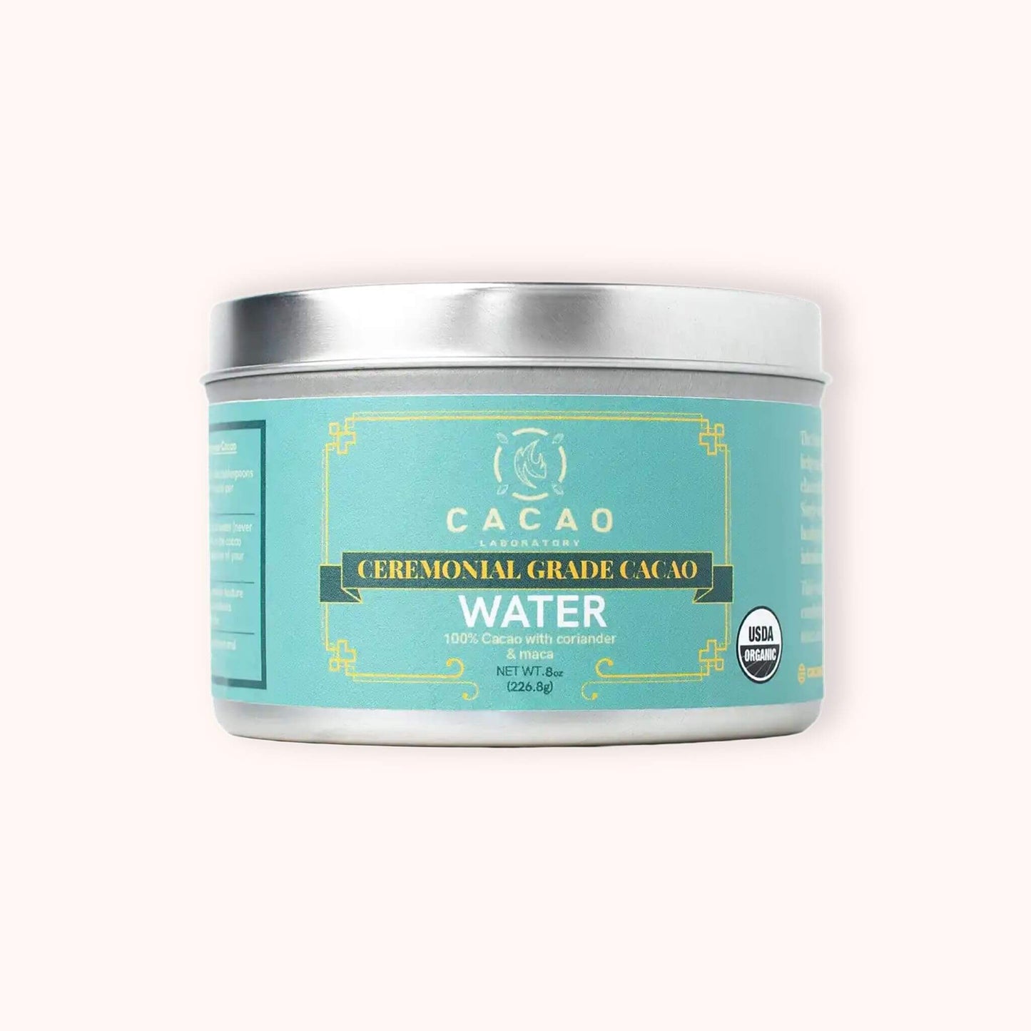 A silver metal tin of Cacao Laboratory Cacao Ceremonial Grade Cacao Water, featuring a light teal label with gold accents and a silver lid, set against a solid pale pink background.