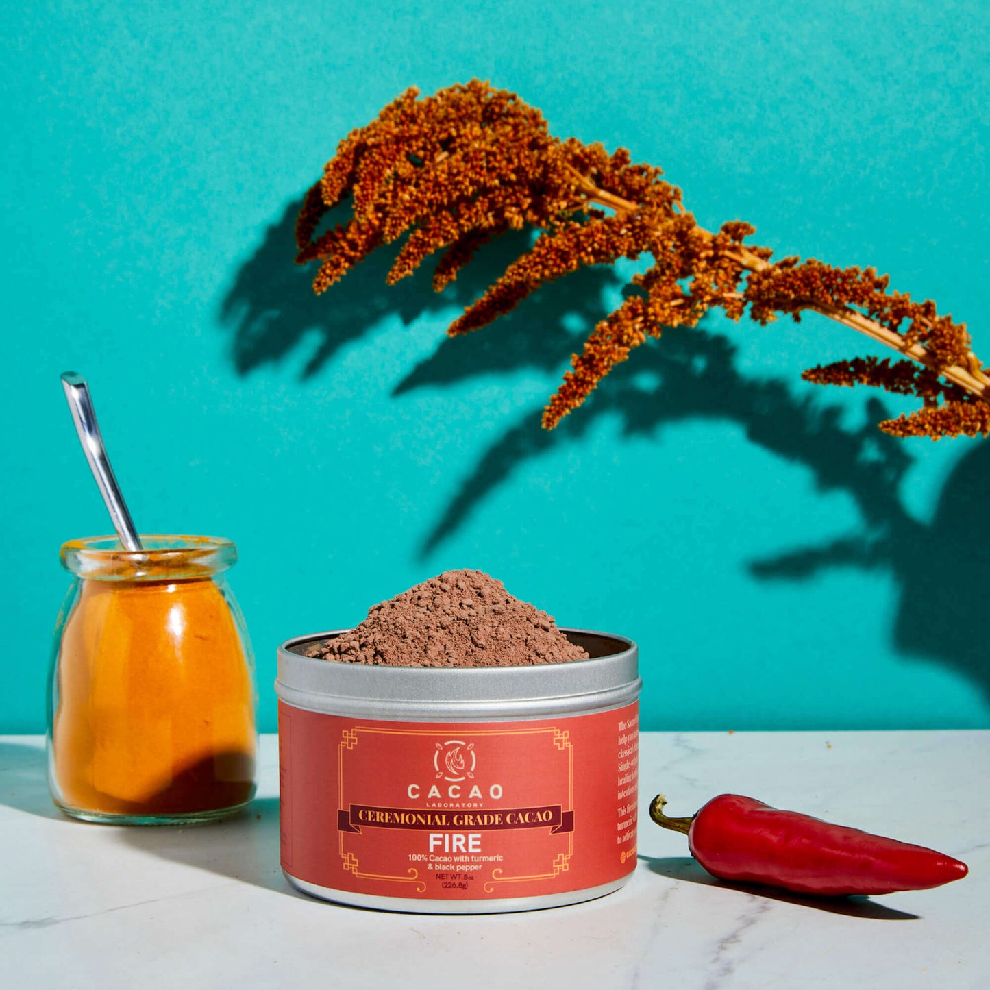 
                  
                    Cacao powder container with a jar of cayenne pepper and a red chili pepper on a teal background
                  
                